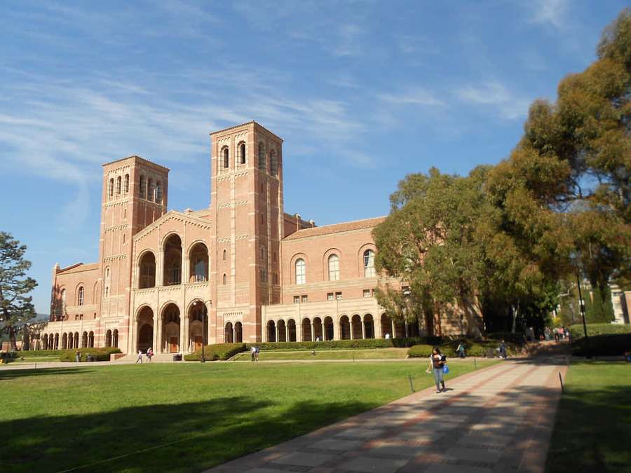 Alunos caminham por um caminho em direção a um histórico edifício universitário de tijolos com torres gêmeas. — Campus da UCLA Papéis de Parede