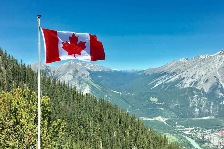 Canadian flag flying high above a mountain valley with forests and snow-capped peaks. — Canada Background