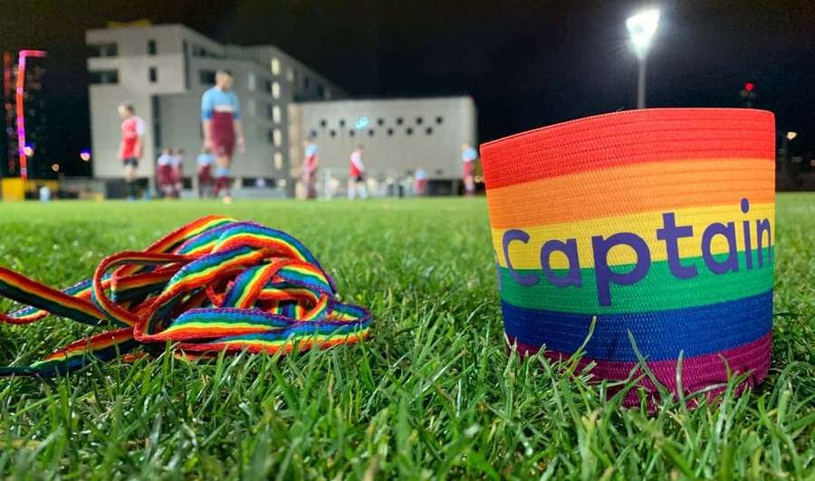 Rainbow captain armband and laces on grass with soccer players in background at night. — Captain Armband Wallpaper