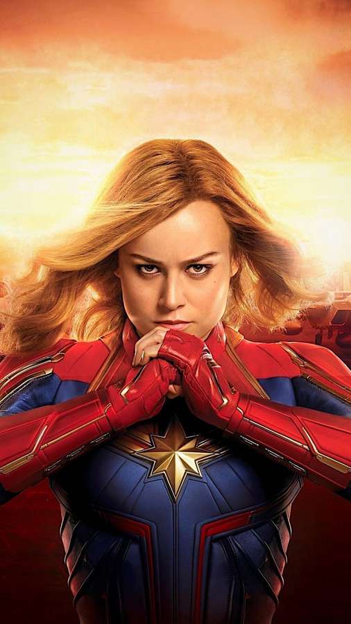 Capitana Marvel en su traje rojo y azul con las manos juntas bajo un cielo de atardecer brillante. — Captain Marvel iPhone Fondos de pantalla