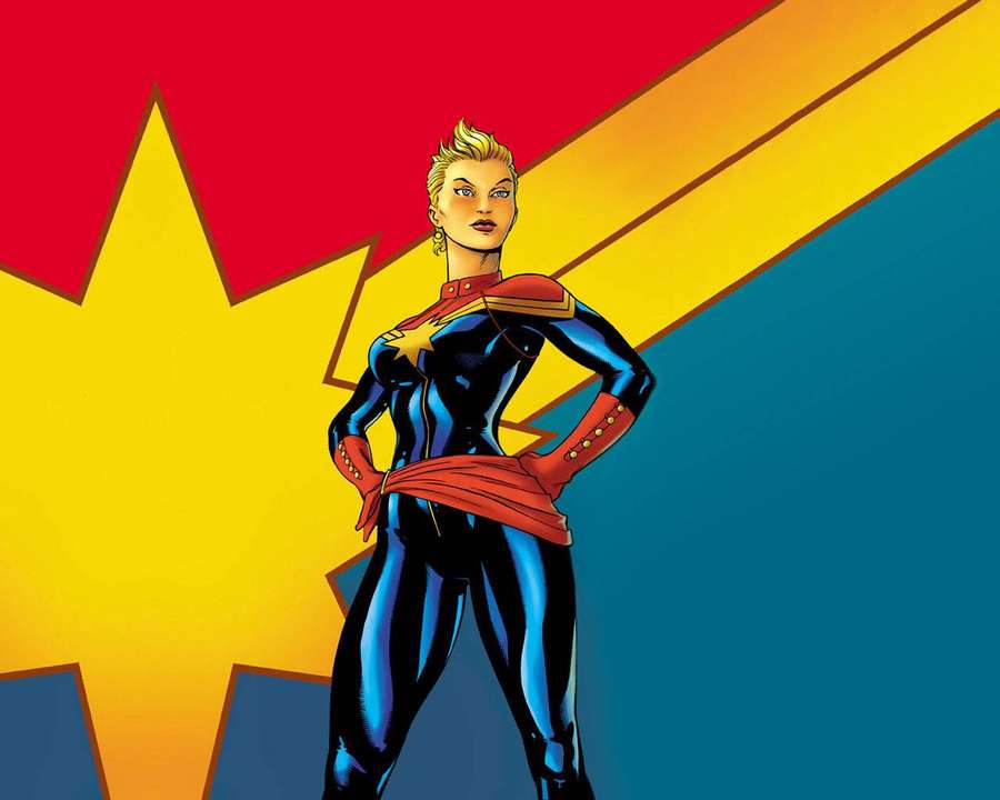 Uma super-heroína em um traje azul e vermelho posa confiantemente contra um fundo de explosão estelar. — Captain Marvel Papéis de Parede