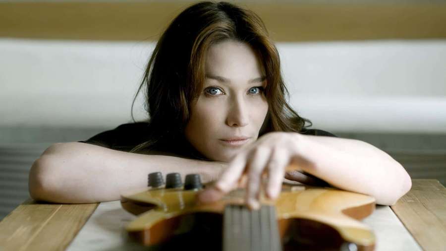 Nainen lepää leukaa käsivarsillaan puisen bassokitaran vieressä pöydällä. — Carla bruni Taustakuvat