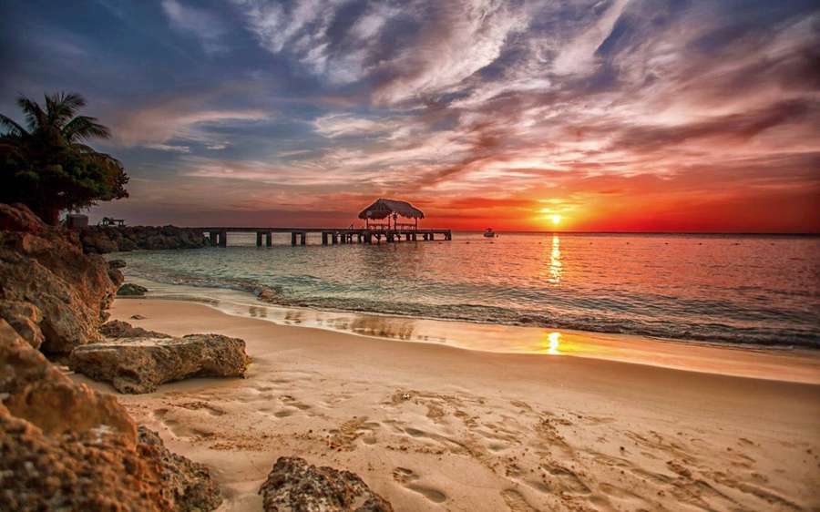 Pôr do sol sobre praia tropical com píer e pegadas — Céu estético Papéis de Parede