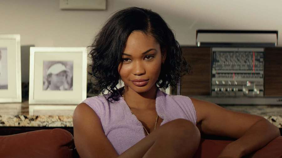 A woman in a purple top sits on a red couch in a room with vintage audio gear and framed photos. — Chanel Iman Wallpaper
