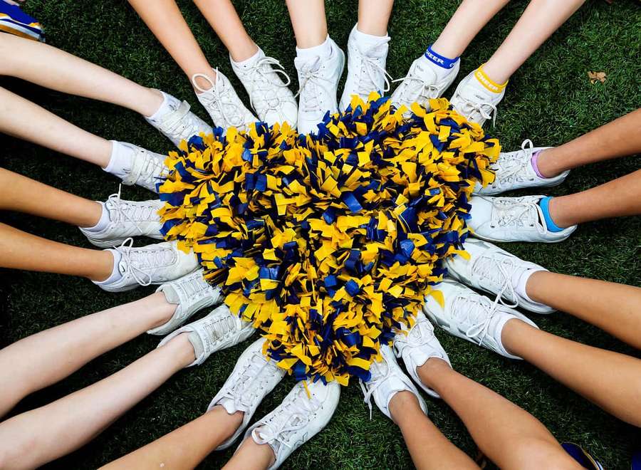Cheerleaders in white sneakers surround a yellow and blue pom-pom on grass. — Cheerleading Aesthetic Wallpaper