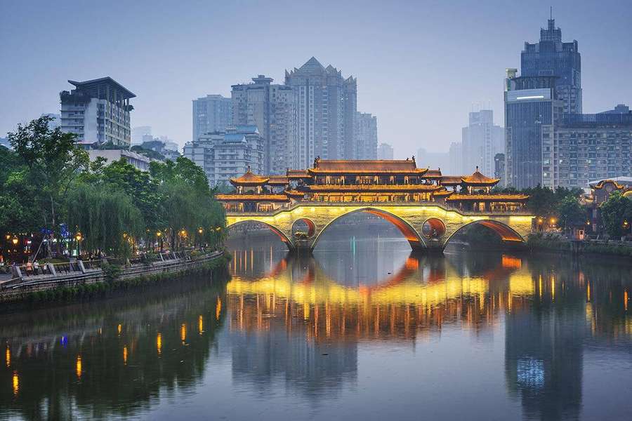 Eine historische chinesische Brücke, die in der Dämmerung beleuchtet ist, mit Spiegelungen im Fluss, umgeben von Stadtgebäuden. — Chengdu Wallpaper