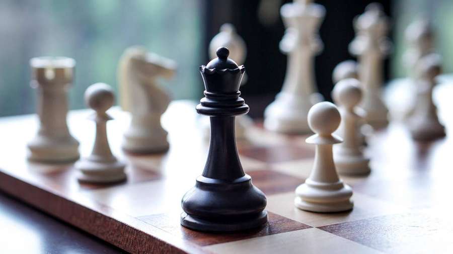 A black chess king on a wooden board with white pieces out of focus behind it. — Chess Wallpaper