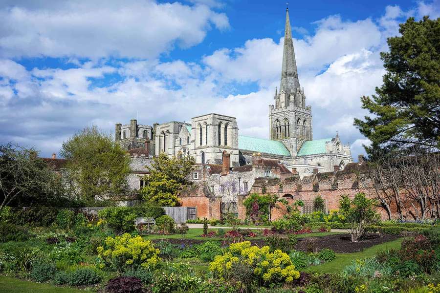 A historic stone cathedral with a tall spire surrounded by a lush garden under a blue sky. — Chichester Wallpaper