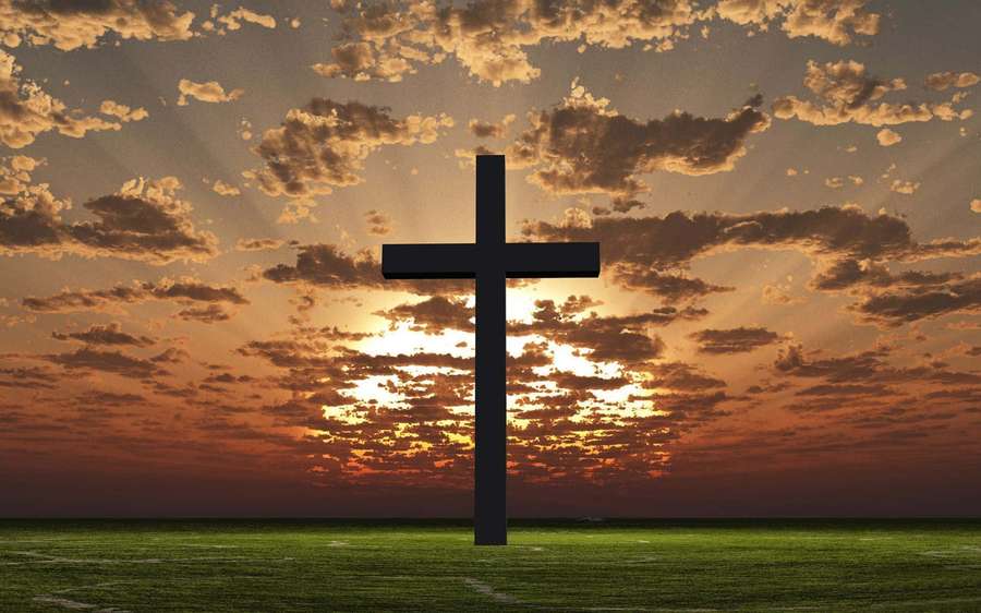 A black cross silhouette on green grass under a vibrant sunset sky. — Christian Background