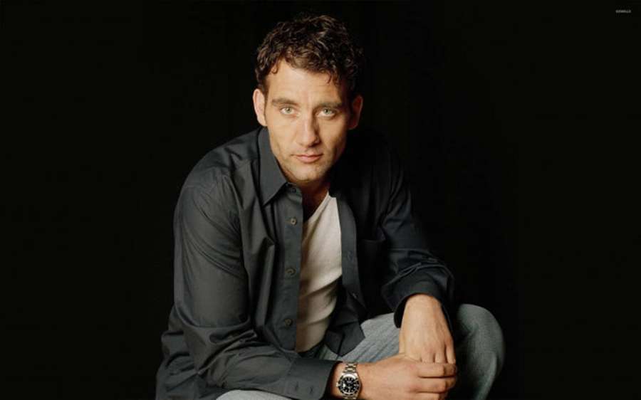 A man in a dark shirt and watch sitting against a black background. — Clive owen Wallpaper