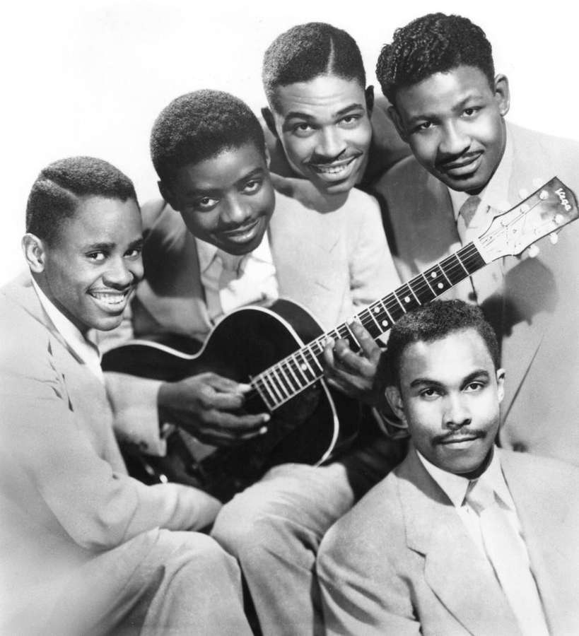 Five men in suits smiling around an acoustic guitar in a studio photo. — Clovers Wallpaper