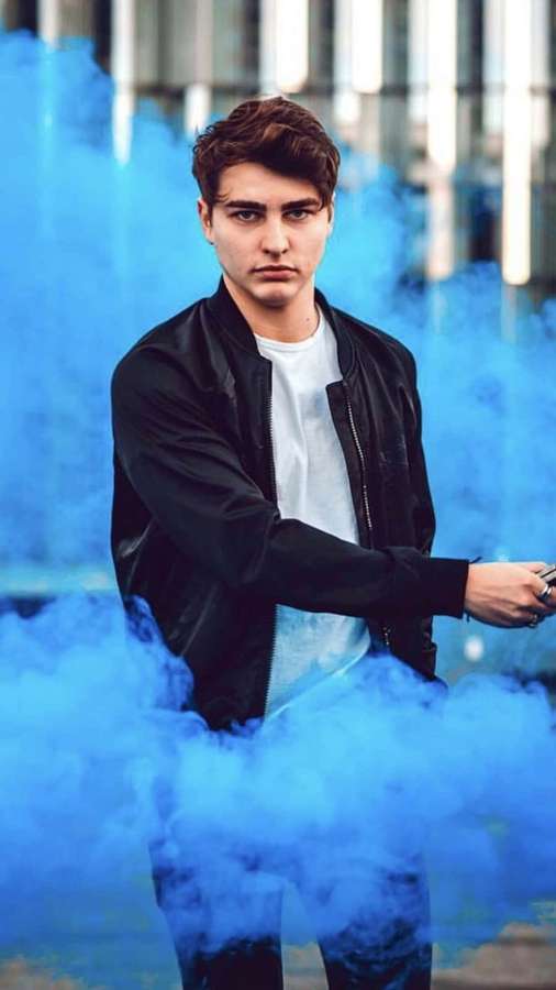 Young man in black jacket surrounded by blue smoke in city setting — Colby Brock Background