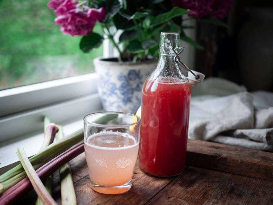 Rhubarb drink in glass and bottle on wooden windowsill with pink flowers. — Cordial Wallpaper
