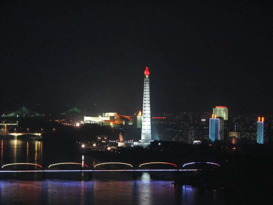 Vista nocturna de un monumento iluminado sobre el agua con un puente y luces de la ciudad. — Corea del Norte Fondos de pantalla