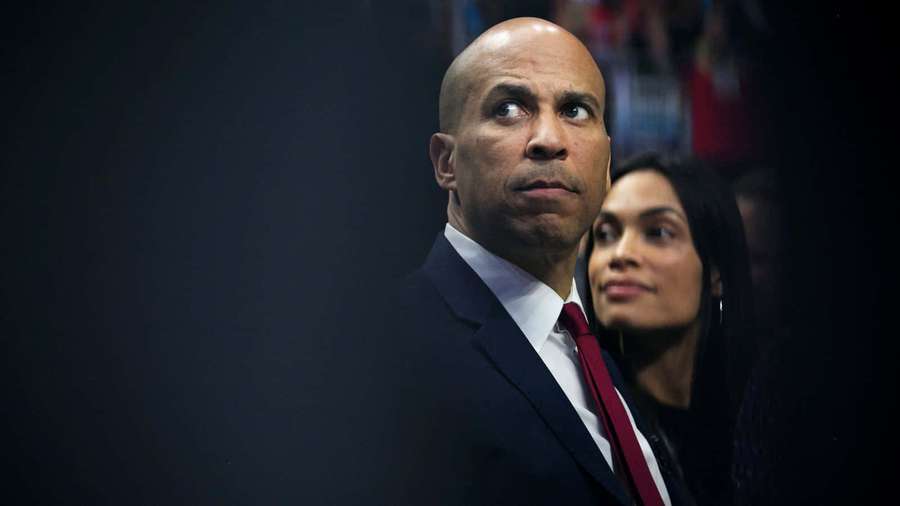 A man in a suit and red tie looks upward with a serious expression while a woman sits beside him. — Cory Booker Wallpaper
