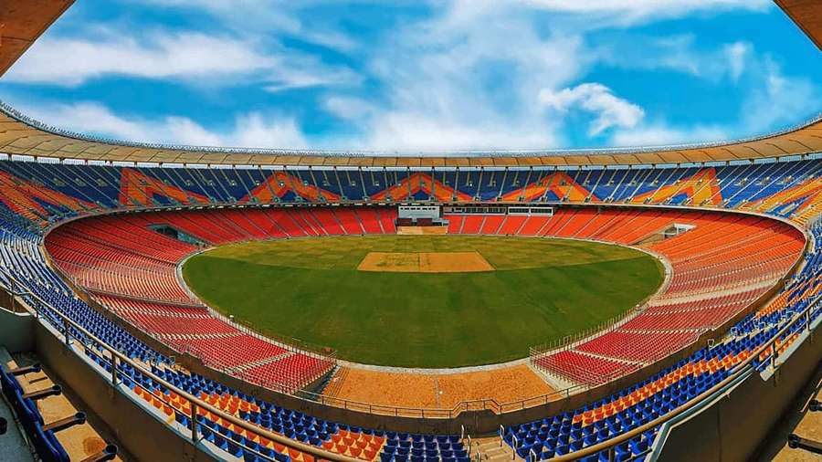 Empty cricket stadium with colorful seats and green field under clear sky. — Cricket Ground Background