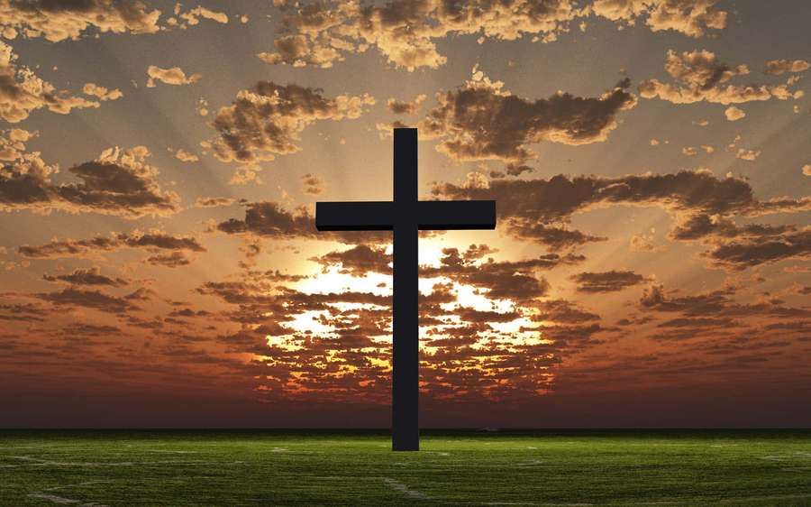A black cross silhouetted against a sunset sky over a green field. — Cross Background