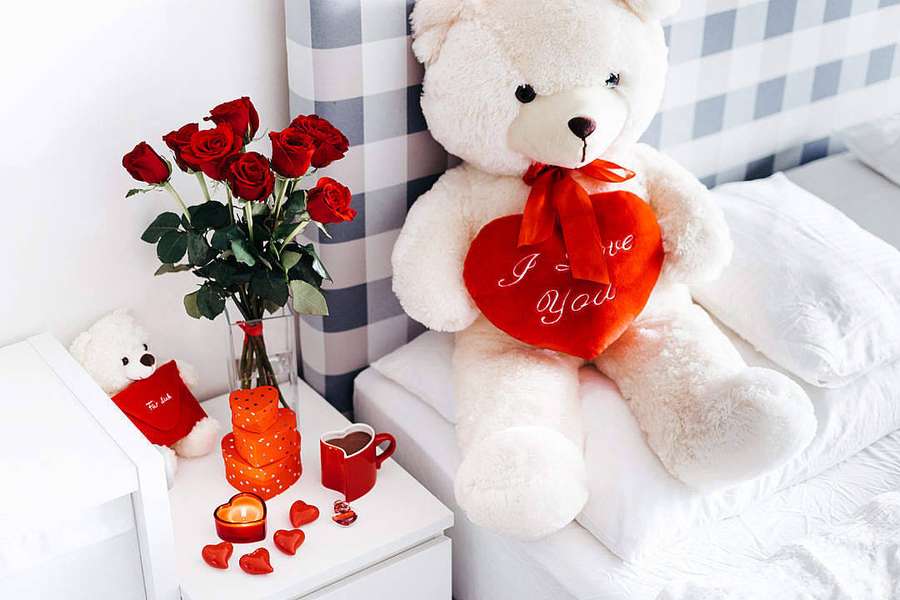 A large teddy bear holding a red heart with I love you text next to red roses and Valentine's decorations on a bed. — Cute Teddy Bear Wallpaper