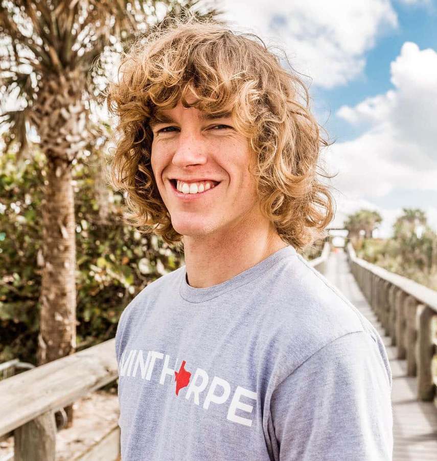 Ung mann med krøllete hår smilende på en solrik promenade med palmer. — Danny Duncan Bakgrunner