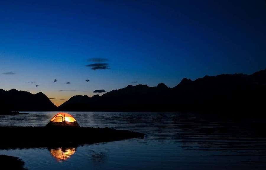 A glowing tent on a lakeshore at twilight with mountains in the background. — Dark Landscape Wallpaper