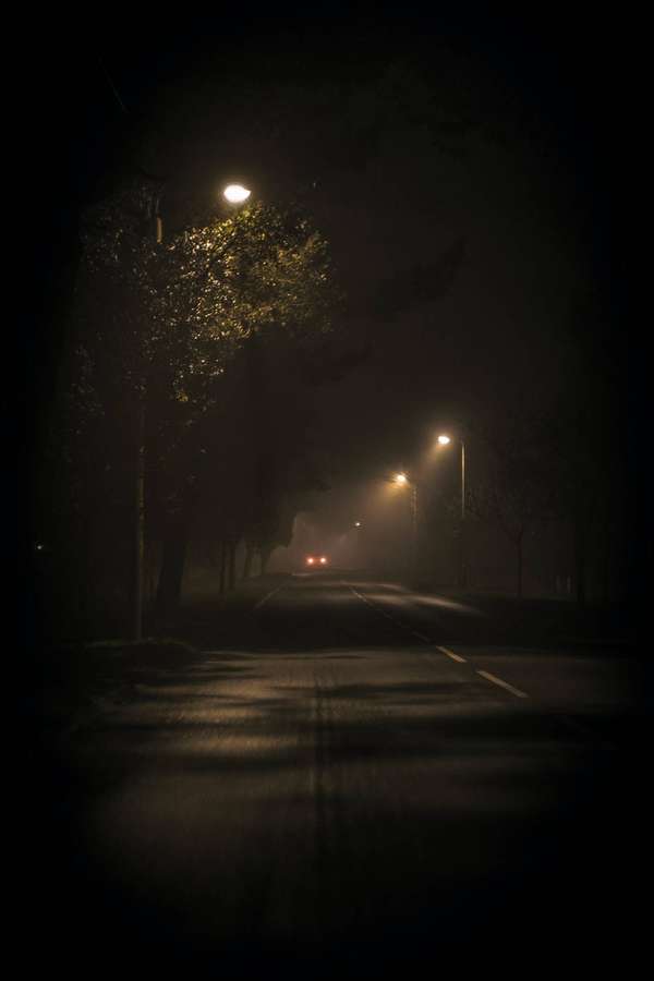 Eine neblige Nachtstraße, beleuchtet von Straßenlaternen, mit den Scheinwerfern eines Autos in der Ferne. — Dark Night Bilder