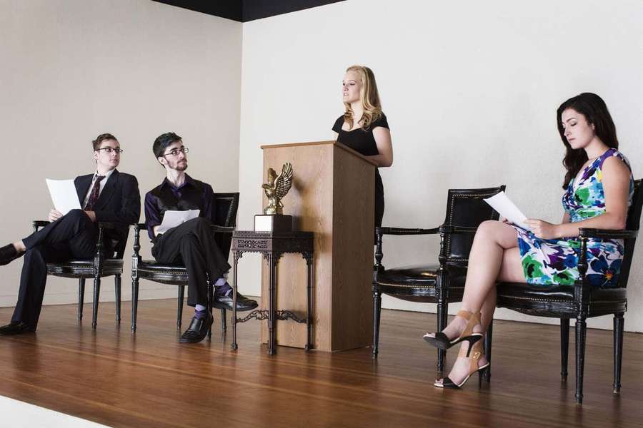 Four people in a modern office setting with one woman speaking at a podium. — Debate Wallpaper
