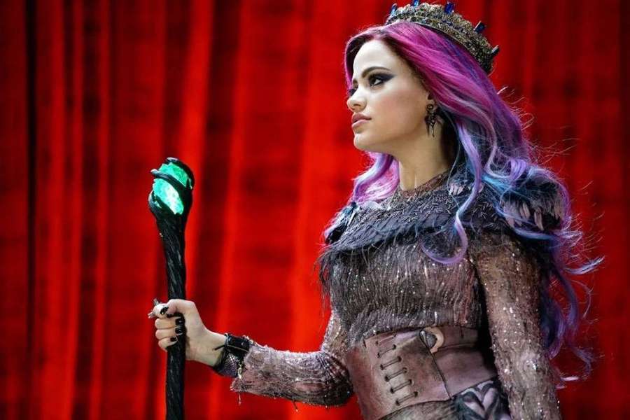 A witch with colorful hair holds a glowing staff on a red stage curtain. — Descendants Wallpaper