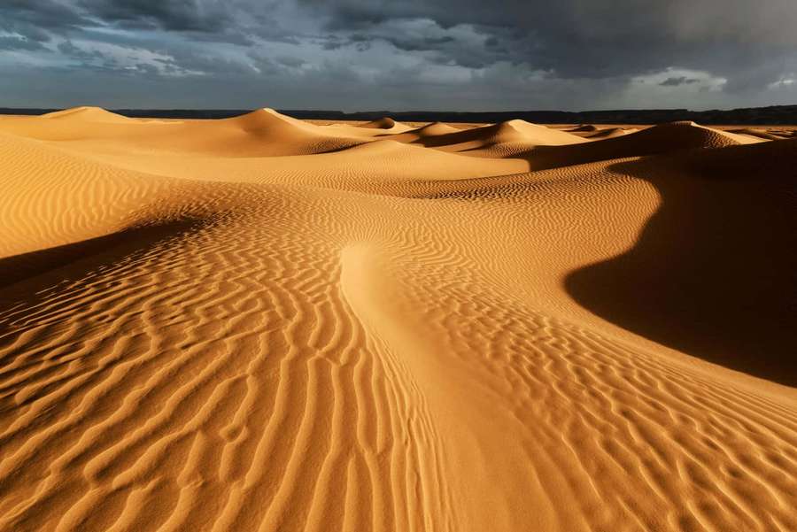 Golden desert dunes under dramatic storm clouds at sunset. — Desert Background