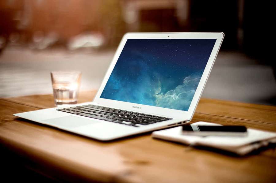 Silver laptop on wooden table with glass of water and notebook nearby. — Desk Background