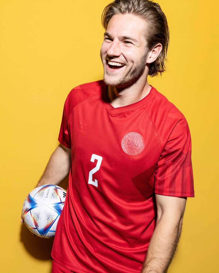 Un sonriente jugador de fútbol masculino con camiseta roja sosteniendo un balón de fútbol sobre fondo amarillo. — Dinamarca Fondos de pantalla