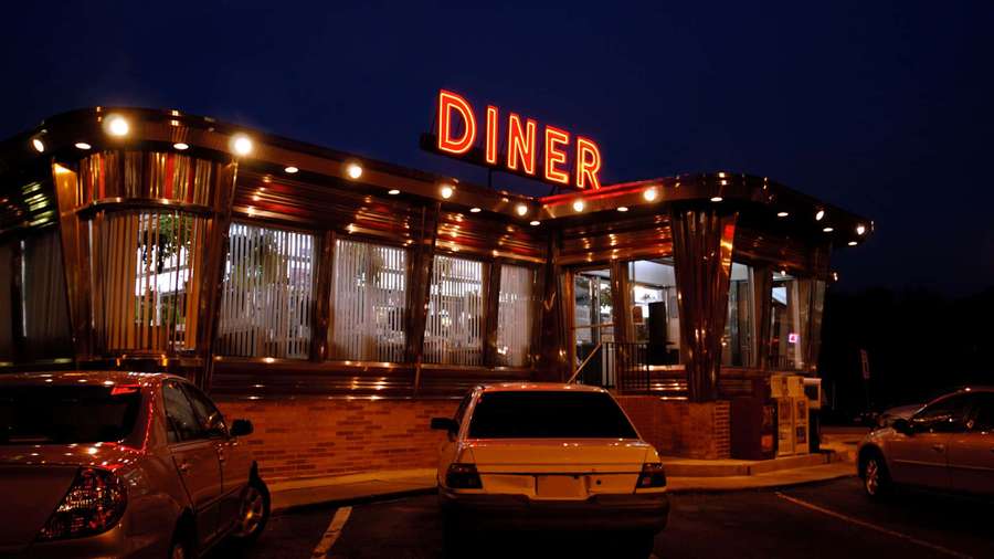A retro diner with a glowing red sign and parked cars at night. — Diner Background