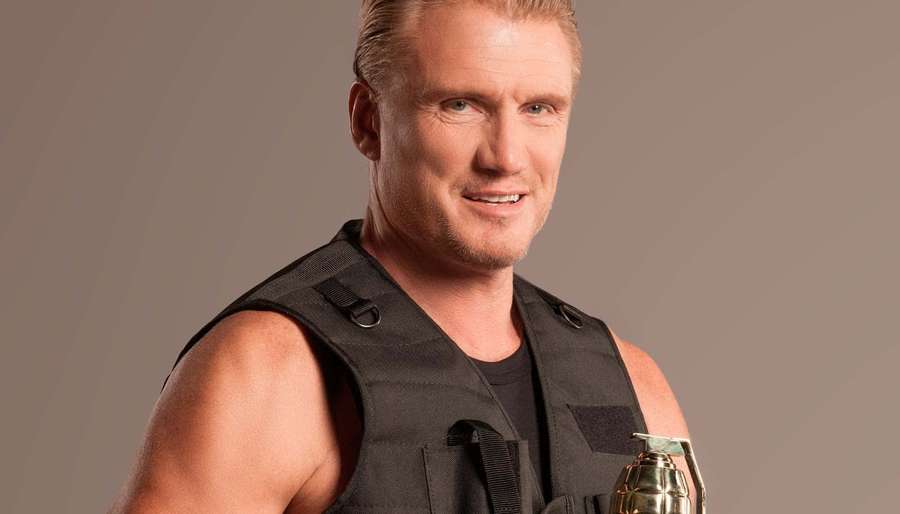 A muscular blond man in a black tactical vest holding a grenade and smiling. — Dolph Lundgren Wallpaper