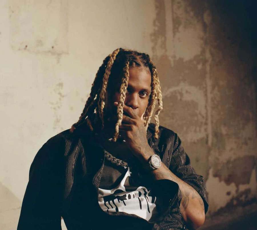 Man with blonde braids and tattoos in a black jacket and skull tee against a weathered wall. — Duh Picture