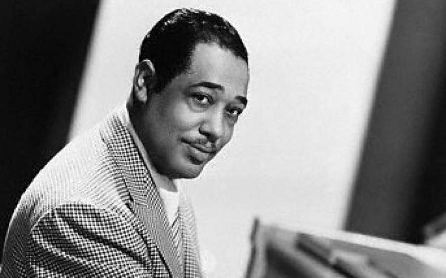 A smiling man in a checkered suit seated at a piano in a studio setting. — Duke Ellington Wallpaper