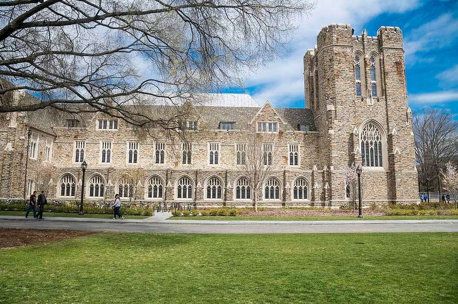 Students walk on a path near a historic stone university building with arched windows. — Durham Wallpaper