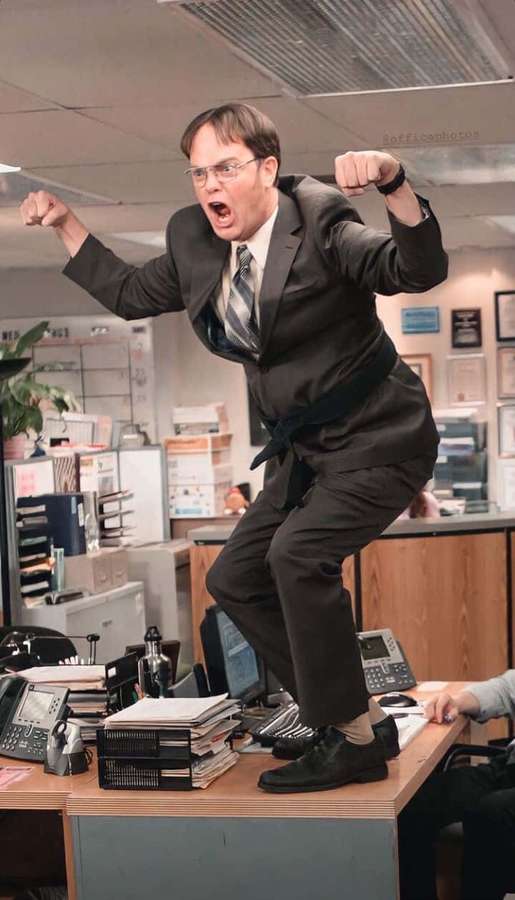 Man in suit jumping on desk in office with fists raised and mouth open. — Dwight schrute Wallpaper