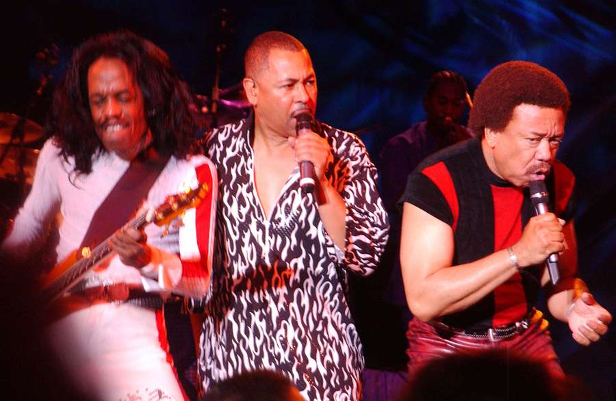 Three musicians performing live on stage with guitars and microphones under colorful lights. — Earth Wind And Fire Wallpaper