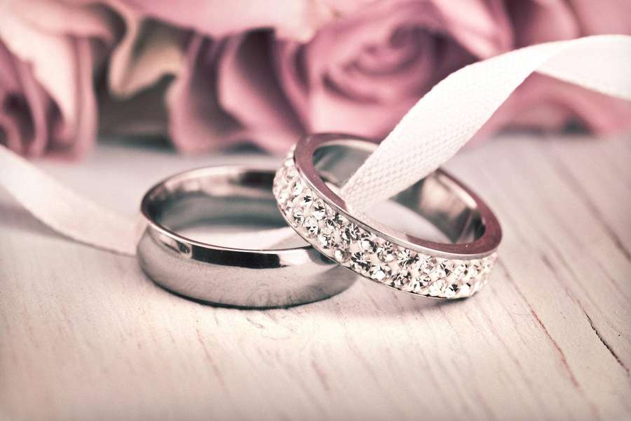Zwei Eheringe, einer schlicht und einer mit Diamanten besetzt, auf einem weißen Band mit rosa Rosen. — Eheringe Wallpaper