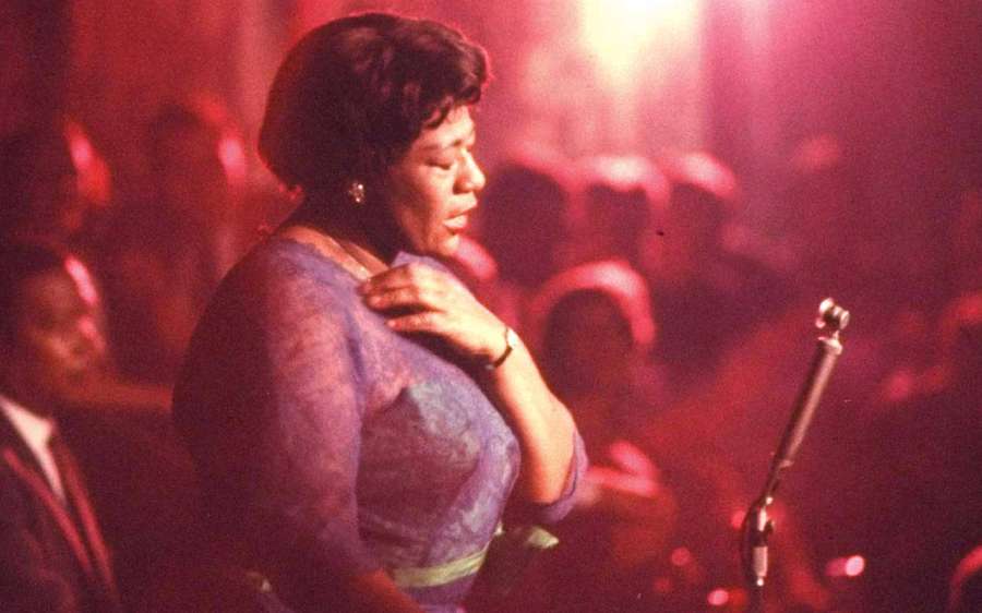 A woman sings into a microphone on stage under red lights with an audience behind her. — Ella Fitzgerald Background