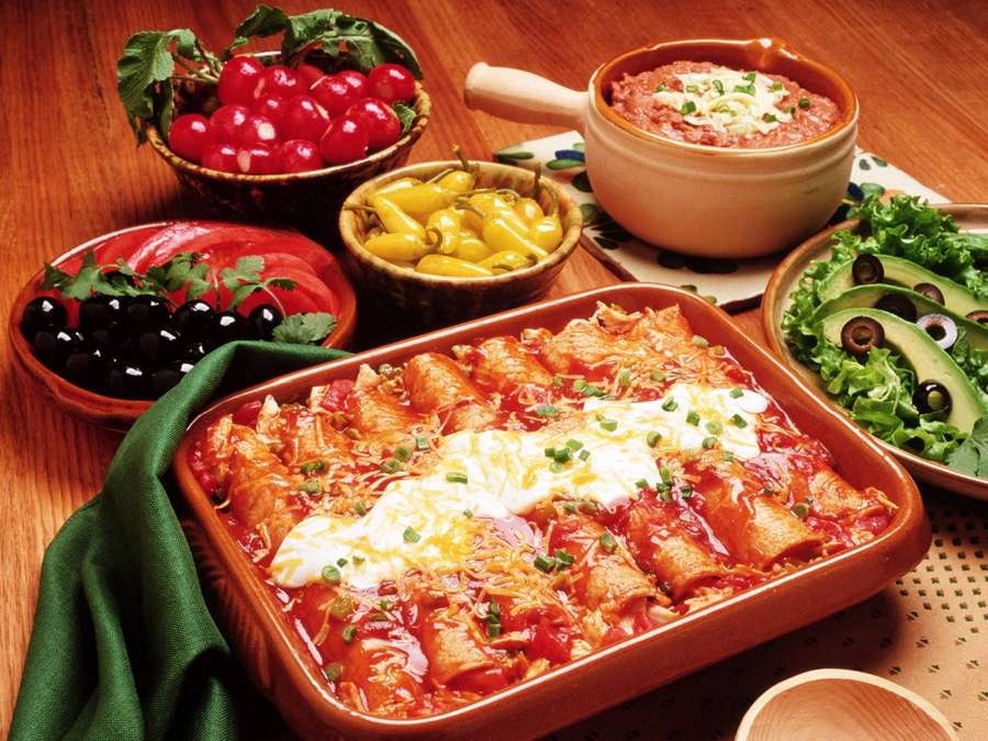 A colorful Mexican dish with enchiladas, avocado, olives, and cherry tomatoes on a wooden table. — Enchiladas Background