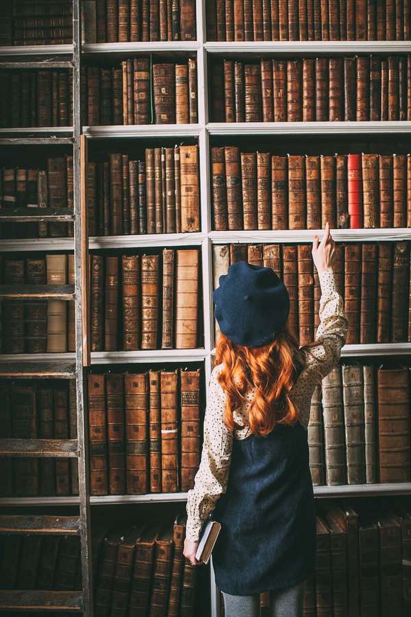 Persona con cabello rojo y boina azul alcanzando un libro rojo en una estantería de madera. — Estantería Fondos de pantalla
