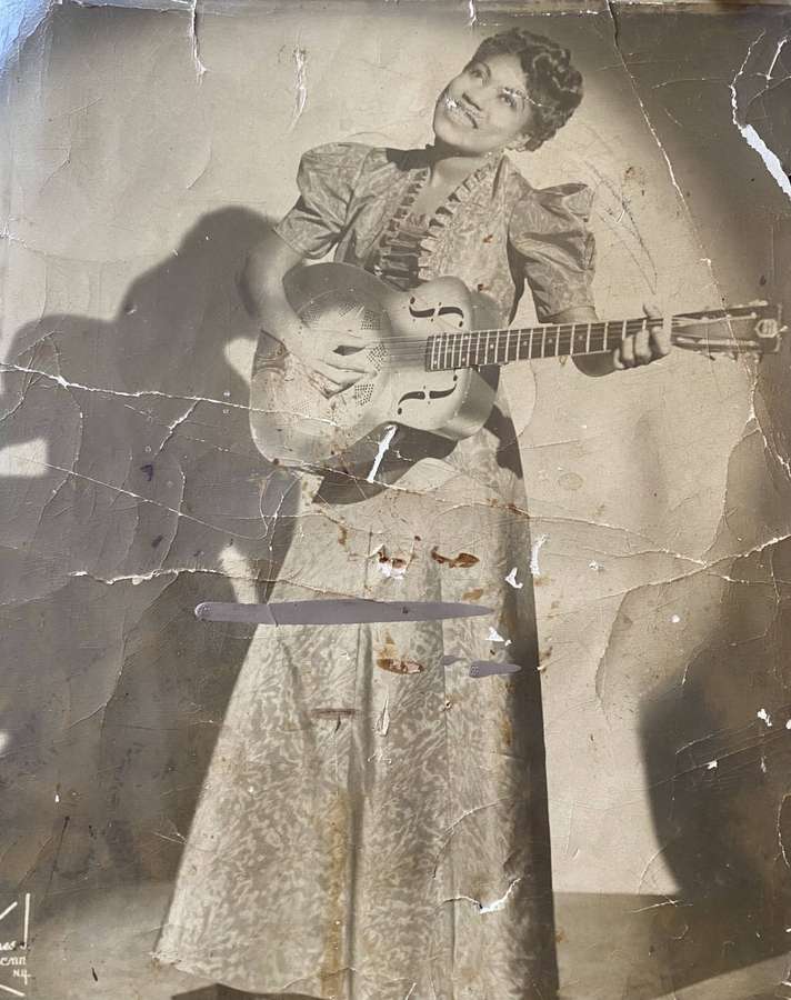 Una mujer sonriente con vestido vintage sosteniendo una guitarra resonadora sobre fondo agrietado. — Evangelio Fondos de pantalla