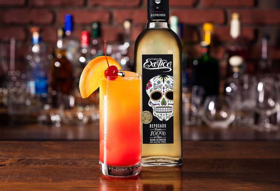 A colorful cocktail with cherry and orange garnish next to a tequila bottle on a bar. — Exotico Wallpaper