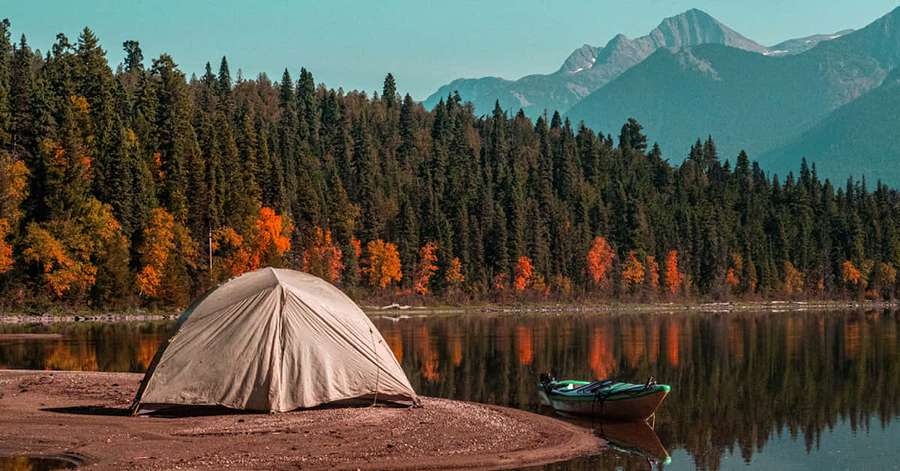 A tent and boat on a calm lake with autumn trees and mountains in the background. — Fall Camping Wallpaper