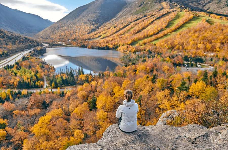 Person sitting on a rock overlooking a lake in autumn foliage. — Fall Hikes Wallpaper