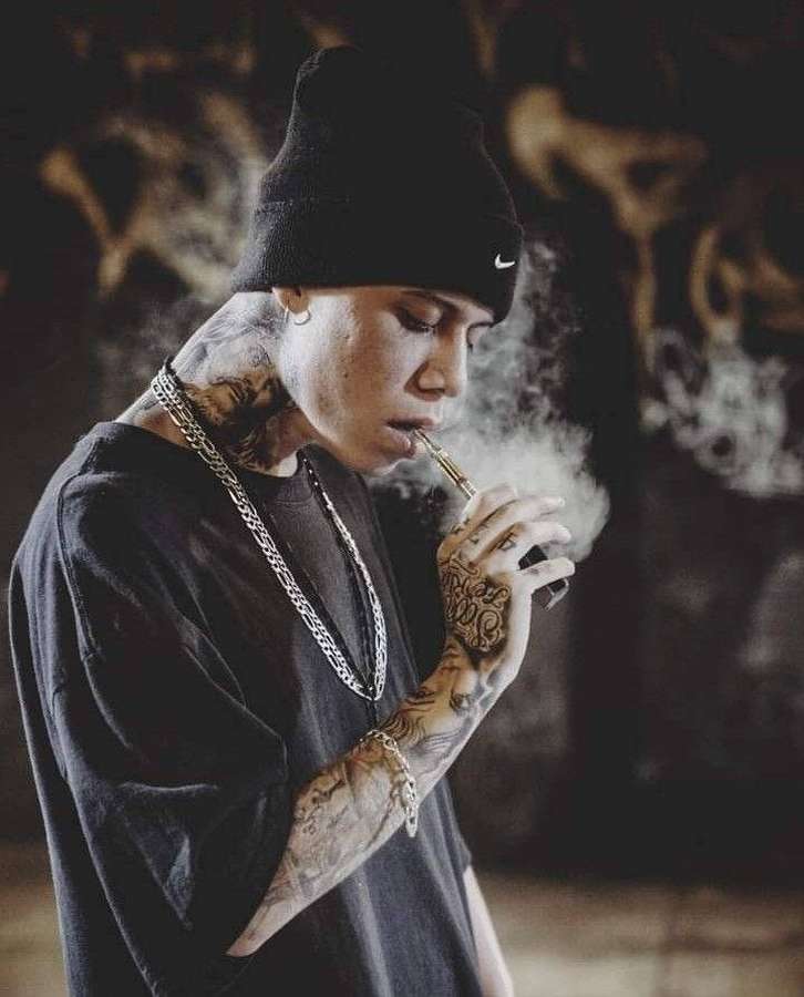 Tattooed person vaping in a graffiti alley wearing a black beanie and chain. — Fe Wallpaper