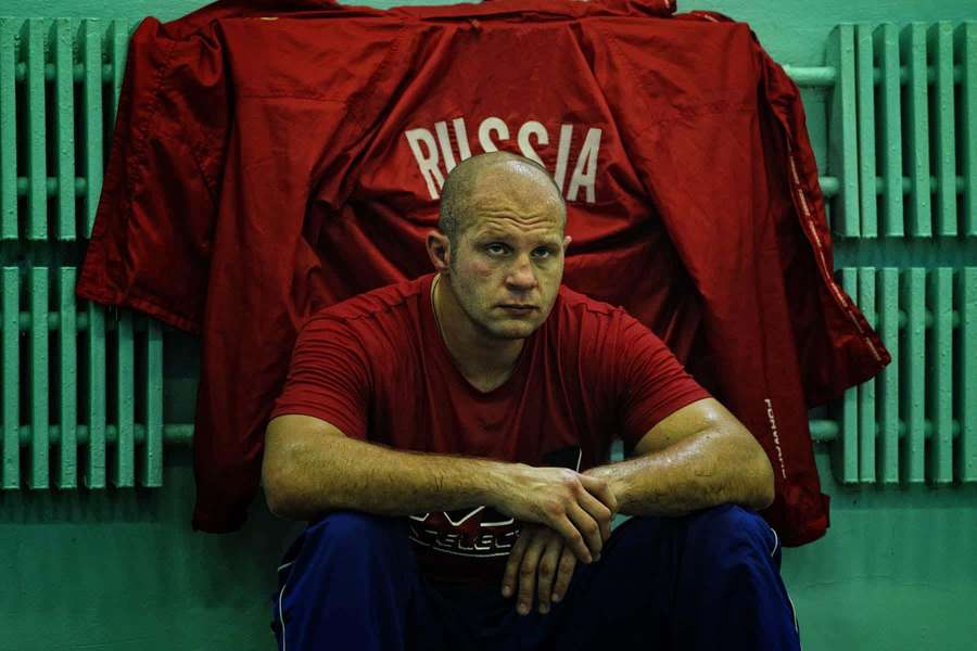 A tired Russian athlete sits in a gym wearing red shirt and blue pants. — Fedor Emelianenko Wallpaper