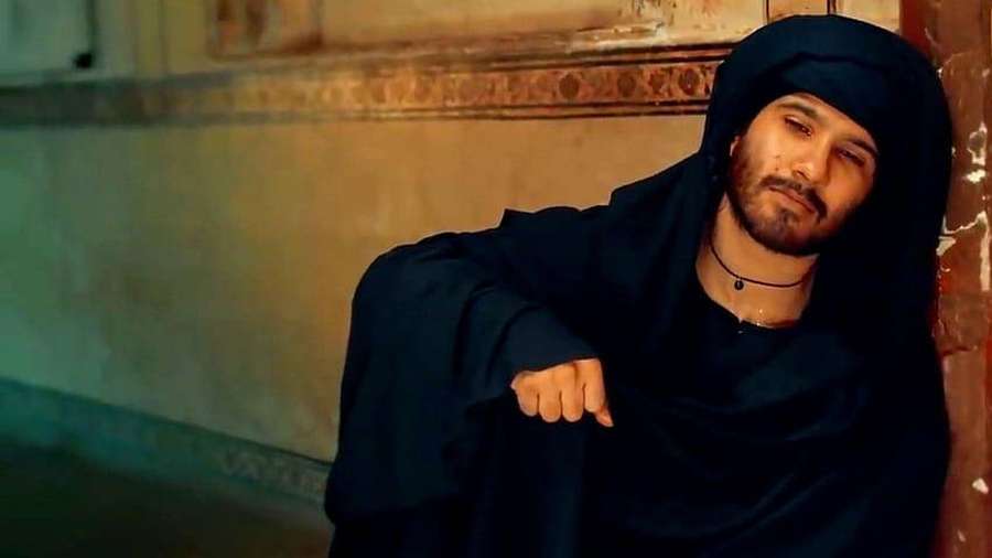 Man in black hooded robe leaning against ornate wall in warm indoor lighting. — Feroz Wallpaper