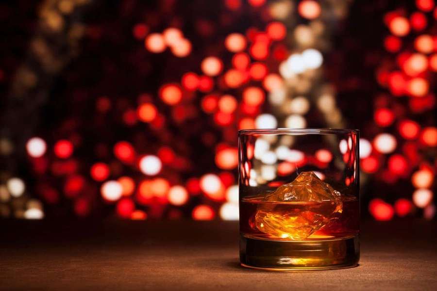 A whiskey glass with ice on a dark surface with red and white lights in the background. — Festive Background