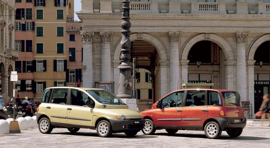 Kaksi kompaktia autoa pysäköitynä aurinkoisella eurooppalaisella kaupungin aukiolla, jossa on historiallisia rakennuksia. — Fiat Multipla Taustakuvat